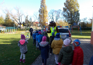 Dzieciaki w ogrodzie przedszkolnym. Podchodzą w rzędzie i witają się kolejno z panem Wyderką - osoba przebrana w strój wydry w kamizelce odblaskowej z logo policji.