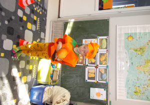 Chłopczyk w pomarańczowym stroju dyni. W rękach trzyma warzywo. Z tyłu na tablicy ilustracje z różnymi gatunkami tego warzywa.
