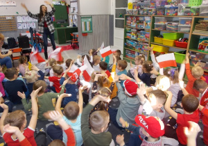 Dzieci siedzą na dywanie. Podnoszą w górę i machają szalikami i flagami.