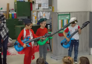 Trzech chłopców stoi przed widownią i gra na dmuchanych elektrycznych gitarach. Na głowach mają wojskowe czapki.