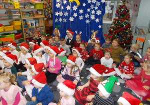 Dzieci siedzą na dywanie i czekają na przybycie Mikołaja. W tyle świąteczna dekoracja: ubrana, kolorowa choinka i niebieskie tło z białymi gwiazdkami.