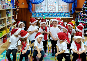 Dzieci siedzą na krzesłach rozstawionych na dywanie. Ubrane są w białe podkoszulki, mikołajowe czapki i czerwone szale. Kolejno mówią swoje role trzymając w ręku mikrofon.