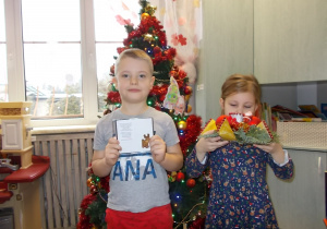 Przy choince stoi dwójka dzeci. Chłopiec trzyma płytę z nagranymi Jasełkami a dziewczynka świąteczny stroik.