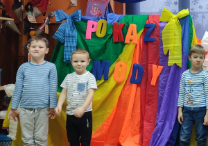Trzech chłopców w bluzkach w paski pozuje do zdjęcia. Za nimi dekoracja: kolorowa chusta z przypiętymi literami tworzącymi napis POKAZ MODY.