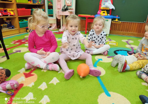 Dzieci siedzą na dywanie w kole. Turlają do siebie pomarańczowy balonik.