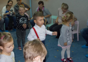 Kilkoro dzieci uśmiecha się, stoi na środku sali i naśladuje ruchy tańca. Z tyłu siedzą rodzice i trzymają swoje dzieci na kolanach.