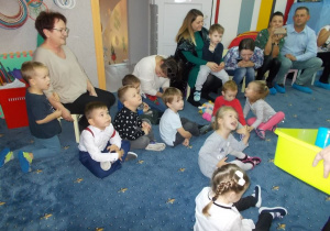 Dzieci siedzą w jednym miejscy na dywanie, twarzą do rodziców. Jedna mama telefonem nagrywa spotkanie.