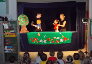Scenografia: na zielonym materiale przymocowane kwiaty tworzące łąkę. Z lewej strony drzewo. Po środku sceny stoją aktorzy trzymając kukiełki chłopca i dziewczynki. Rozmawiają ze sobą.