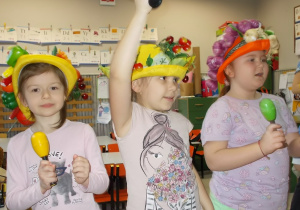 Trzy dziewczynki. Na głowach mają kapelusze ozdobione wiankami z plastikowych owoców i warzyw. W rękach trzymają grzechotki.