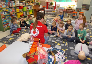 Dzieci siedzą przed wolontariuszką na dywanie. Słuchają pogadanki.