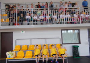 Czwórka chłopców w żółtych koszulkach i granatowych spodenkach siedzi na krzesłach przewidzianych dla zawodników. Nad nimi widownia i pozostałe dzieci z grupy.