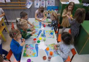 Dzieci siedzą przy dugim stole. Przed każdym podkładka oraz kubeczek z ciastoliną.