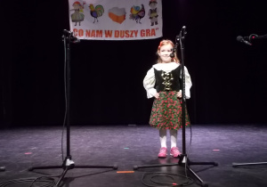 Dziewczynka stoi na scenie przed mikrofonem. Ma kwiecistą spódniczkę. Bluzkę z falbankami i czarną kamizelkę ozdobioną cekinami.