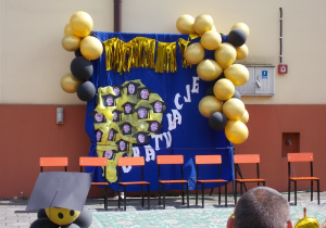 Dekoracja przed budynkiem przedszkola. Rozwieszony niebieski materiał. Po bokach girlanda z złoto-czarnych balonów oraz kwiaty. Na niebieskim tle zdjęcia dzieci kończących przedszkole.