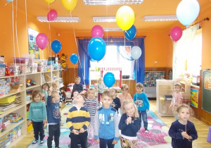 Dzieci stoją na dywanie w różnych częściach sali. Uśmiechają się. Każde trzyma za tasiemkę balonik wiszący pod sufitem.