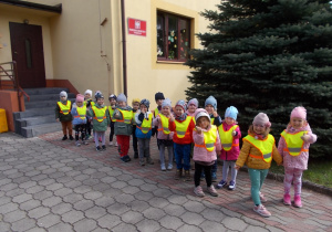 Dzieci przygotowane do spaceru. Ustawione na chodniku przed przedszkolem para za parą. Każde ma na sobie kamizelkę odblaskową.