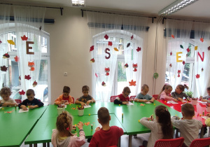 Dzieci siedzą przy długim stole. Każde nakleja kolorowe, papierowe listki na swoje drzewo narysowane na kartonie.