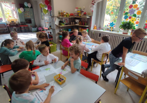 Dzieci siedzą przy stolikach. Kredkami świecowymi kolorują misia z balonikami.