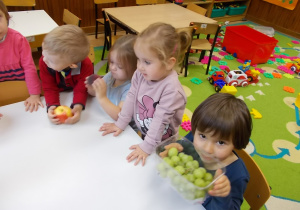 Grupka dzieci stoi przy stoliku. Jedna dziewczynka trzyma pojemnik z winogronem.