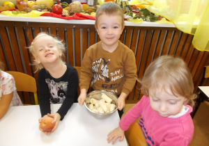 Grupka dzieci stoi przy stoliku. Chłopczyk trzyma miskę z obranymi bananami.