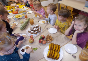 Dzieci siedzą przy długim stole. Każde ma talerzy. Przed nimi zdrowe ciasta, ciasteczka i bułeczki przygotowane przez rodziców.