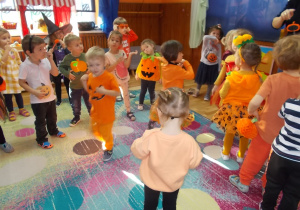 Dzieci tańczą na dywanie. Niektórzy w pomarańczowych ubrankach lub gotowych opaskach i strojach dyni.