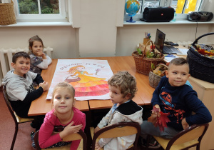 Grupka dzieci siedzi w szkolnej ławce. Przed nimi plakat Pani Jesieni do ozdobienia.