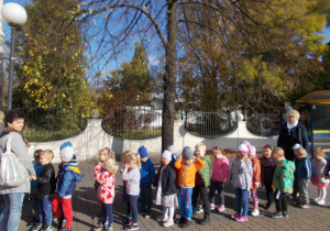 Dzieci ustawione para za parą. Za nimi ozdobny mur parku i kolorowe jesienne drzewa.