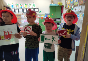 Czterech chłopców w czerwonych kaskach na głowie. Każdy trzyma obrazek symbolizujący różne zachowania bezpieczne.