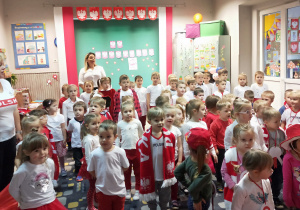 Dzieci stoją na baczność i śiewają hymn. Każdy ma element stroju w kolorze białym i czerwonym. Widać czapki, szaliki, kotyliony, itp.