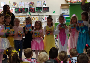 Dziewięć dziewczynek w stroju księżniczek ustawionych w szeregu. Każda trzyma książeczkę z bajkami.