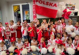 Wszystkie przedszkolaki w strojach biało-czerwonych pozują do zdjęcia. Nad nimi zawieszona flaga Polski i rozciągnięty szalik kibica.
