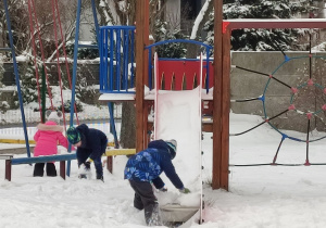 Dzieci toczą kule ze śniegu zgarniętego ze zjeźdżalni i huśtawek.
