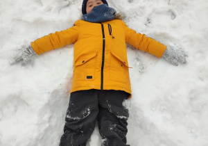 Chłopiec w żółtej kurtce leży na śniegu z rękoma w bok.