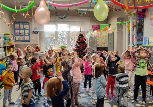 Sala udekorowana w zwisające balony i serpentyny. Dzieci tańczą w rozsypce na dywanie.
