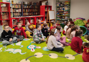 Dzieci siedzą na pufach i dywanie. Za nimi regały z książkami.
