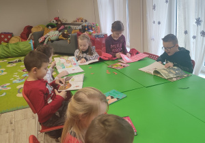 Dzieci siedzą przy długim stole i oglądają książeczki.