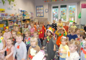 Dzieci stoją na dywanie. Ubrania mają w kolorach jesiennych. Niektórzy przymocowane do ubrań listki lub kapelusze na głowach.