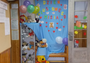 Ścianka. Na niebieskim tle napis Dzień Pluszowego Misia. Obok szafka a na półkach poustawiane różne pluszowe misie. Na góze kolorowe balony.