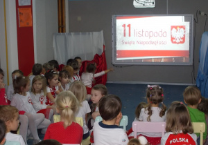 Dzieci siedzą na krzesłach. Z lewej strony dzieci w strojach biało-czerwonych. Po środku, na tablicy napis 11 listopada Święto Niepodległości.