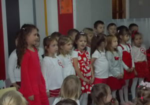 Dzieci stoją i śpiewają piosenki patriotyczne. W pierwszym rzędzie dziewczynki, za nimi chłopcy.