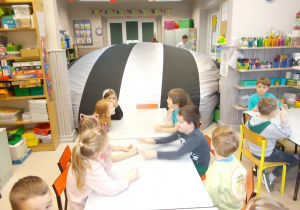 Dzieci siedzą przy stolikach. W drugiej części sali rozkłada się duża kopuła planetarium.