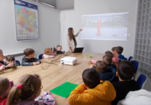 Dzieci siedzą w sali konferencyjnej. Przy rzutniku mama Wojtka objaśniająca pojawiające się kolejne slajdy.