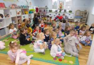 Dzieci siedzą na dywanie, trzymają swoje pluszaki i oglądają jedną z bajek, w której bohaterem jest miś.