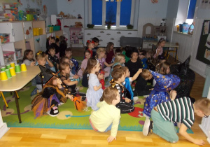Dzieci siedzą na dywanie w przyciemnionej sali. Oglądają prezentację na temat tradycji wróżb andrzejkowych.