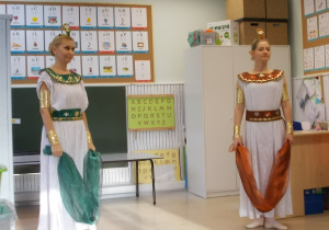 Dwie panie w białych sukniach w stylu greckim. Na głowach mają diademy a w rękach szarfę.