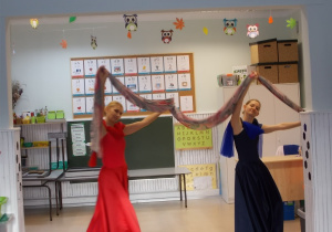 Dwie panie w czerwonej i niebieskiej sukience. Nad głową wspólnie trzymają długi materiałowy szal.