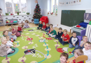 dzą w rzędach na dywanie. Z lewej strony dziewczynki z prawej chłopcy. W głębi ubrana choinka a obok przyozdobione czerwonym materiałem miejsce dla Mikołaja.