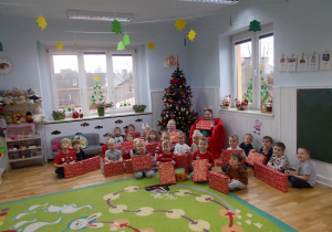 Zdjęcie grupowe. Dzieci siedzą przed choinką, trzymają pięknie zapkowane w czerwony papier prezenty.