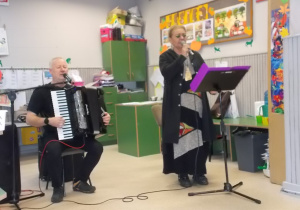 Pan siedzi i gra na akordeonie. Pani stoi przy mikrofonie. Oboje śpiewają.
